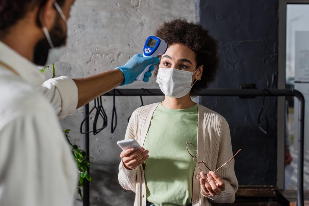 Αφροαμερικανός πελάτης με ιατρική μάσκα κρατώντας smartphone κοντά σε σερβιτόρο με υπέρυθρο θερμόμετρο σε θολή πρώτο πλάνο  - Φωτογραφία, εικόνα