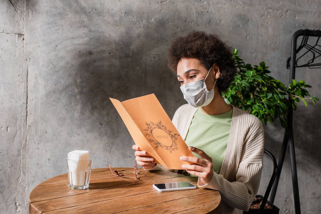 Африканская американка в медицинской маске рассматривает меню возле смартфона в кафе  - Фото, изображение