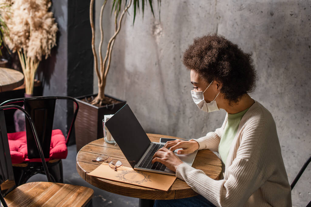 Freelancer afroamericano en máscara médica usando laptop cerca del menú en la cafetería  - Foto, Imagen