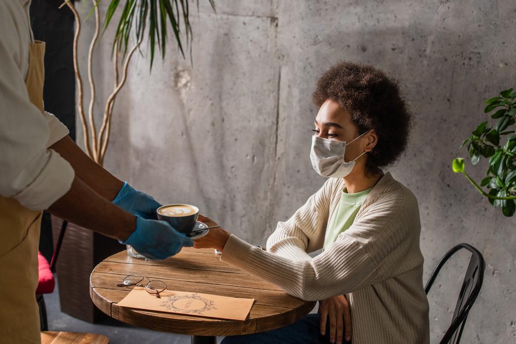 Африканский американский официант в латексных перчатках держит кофе рядом с женщиной в медицинской маске в кафе  - Фото, изображение