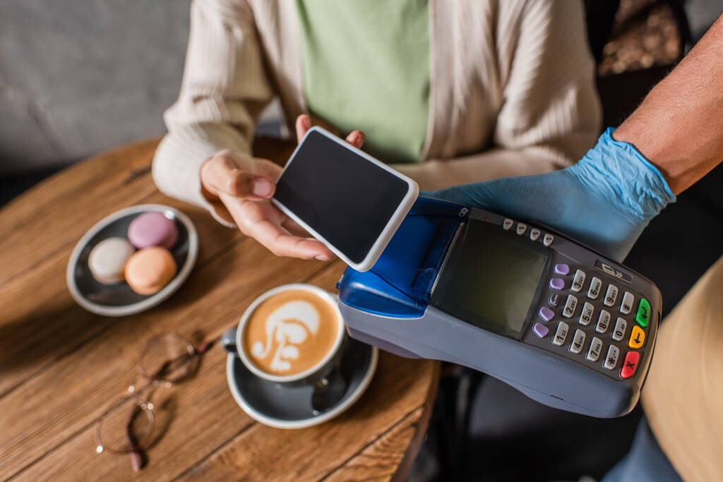 Vue recadrée d'un client flou payant avec un smartphone près d'un serveur afro-américain en gant de latex avec terminal de paiement  - Photo, image