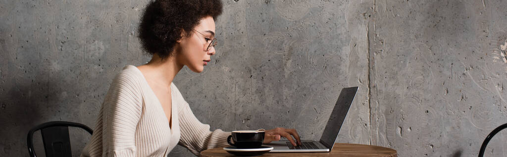 Vista lateral del freelancer afroamericano utilizando el ordenador portátil cerca de taza en la cafetería, pancarta  - Foto, Imagen