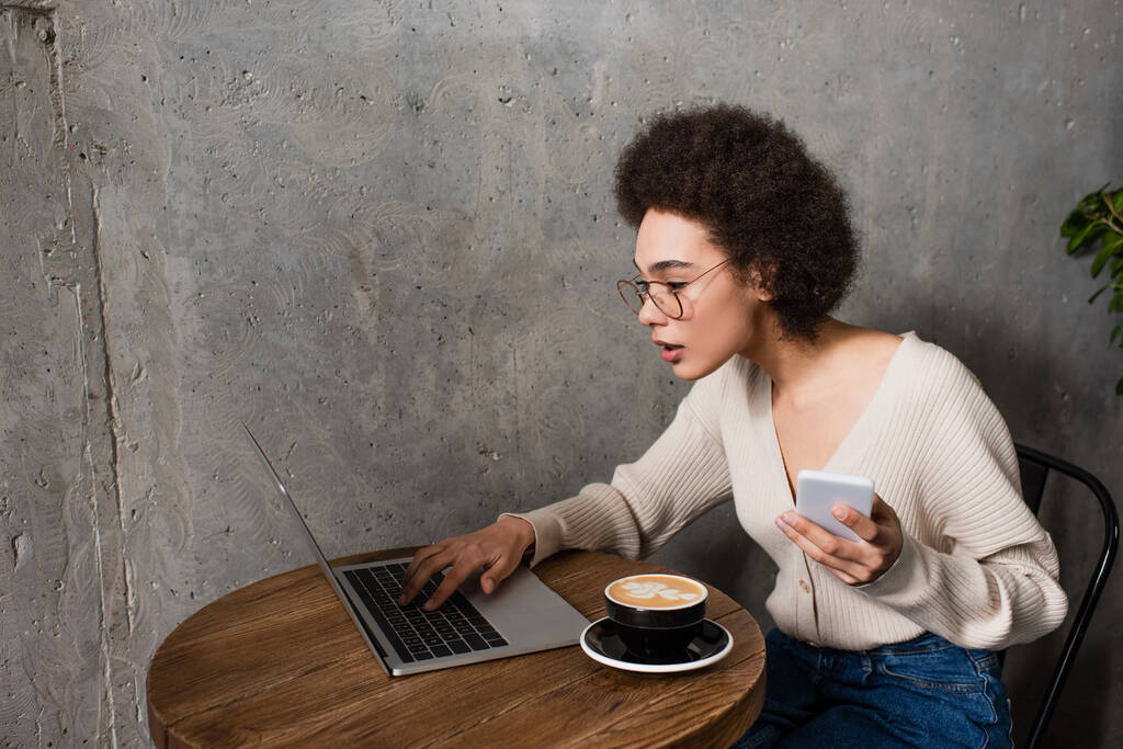 Afrikai amerikai teleworker okostelefonnal és laptoppal, a cafe cappuccino közelében dolgozik  - Fotó, kép
