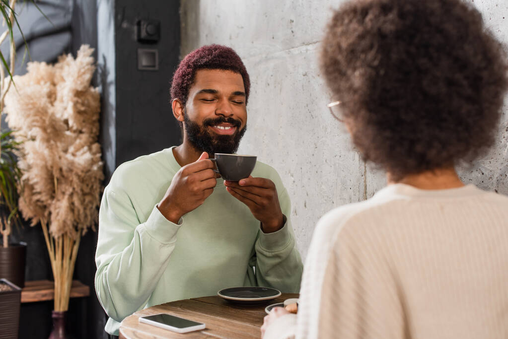 Uśmiechnięty Afroamerykanin z kawą siedzący obok zamazanej dziewczyny w kawiarni  - Zdjęcie, obraz