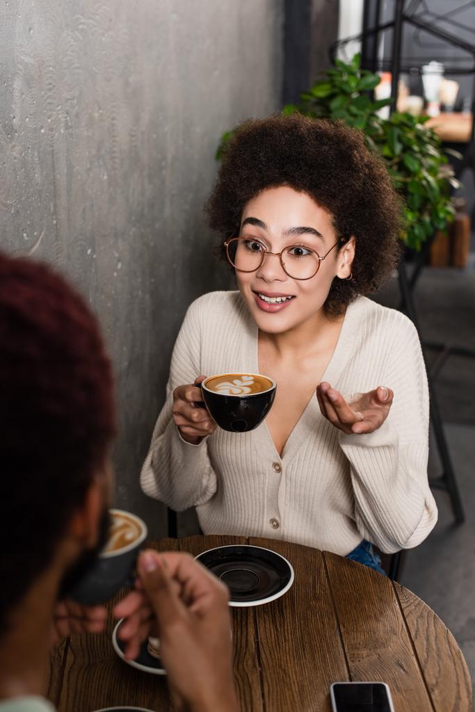 glimlachende Afrikaan amerikaanse vrouw met koffie praten met wazig vriendje in cafe  - Foto, afbeelding