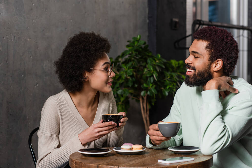 Χαμογελώντας αφροαμερικάνικο ζευγάρι με καφέ μιλώντας κοντά σε αμυγδαλωτά στο καφέ  - Φωτογραφία, εικόνα