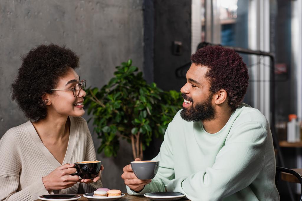 Vista lateral de la sonriente pareja afroamericana sosteniendo café cerca de macarrones en la cafetería  - Foto, Imagen