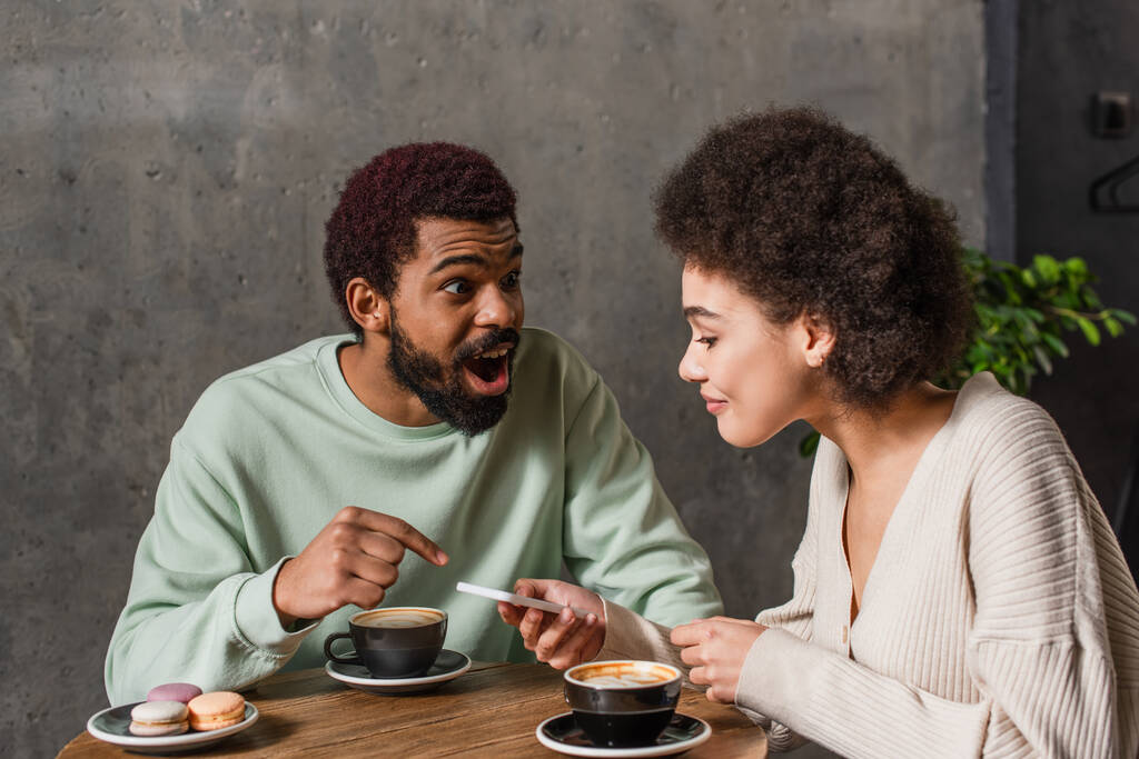 Heyecanlı Afrikalı Amerikalı adam, kafeteryadaki akıllı telefon ve kahveyle sevgilisini işaret ediyor.  - Fotoğraf, Görsel