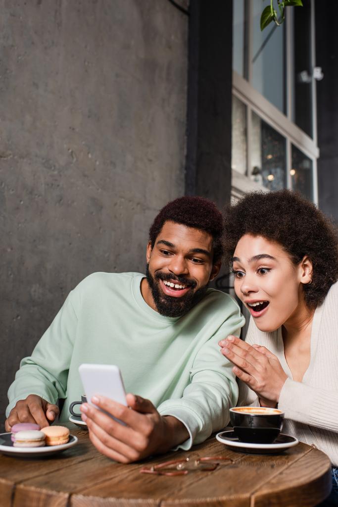 興奮したアフリカ系アメリカ人のカップルは、カフェでマカロンとコーヒーの近くのスマートフォンを使用して  - 写真・画像