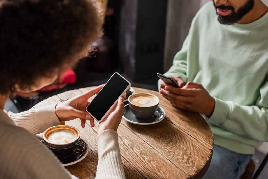 Smartphone con pantalla en blanco en manos de mujer afroamericana sentada cerca de novio y café en cafetería  - Foto, Imagen