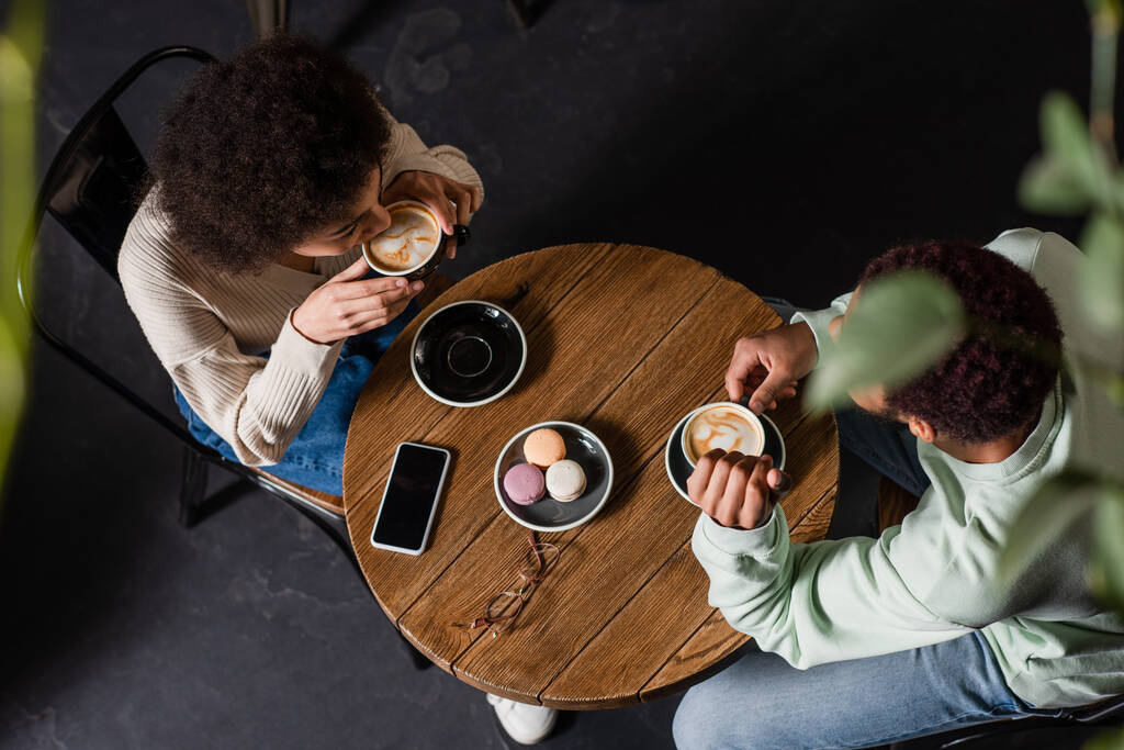 Vista aérea de una joven pareja afroamericana tomando café cerca de macarrones y teléfonos inteligentes en la cafetería  - Foto, imagen