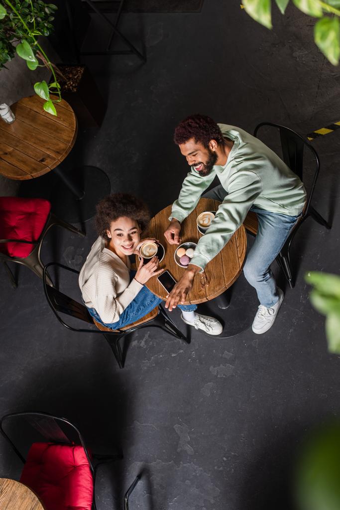 Вид зверху усміхнений афроамериканський чоловік махає рукою біля дівчини з кавою в кафе
  - Фото, зображення