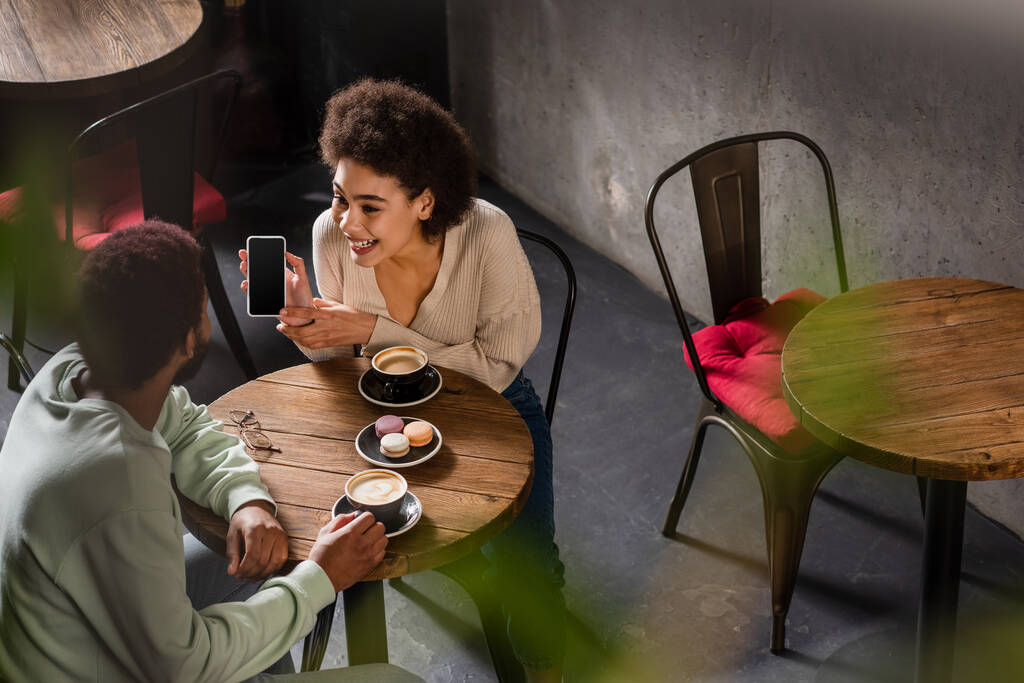 Femme afro-américaine joyeuse montrant smartphone au petit ami avec café dans le café  - Photo, image