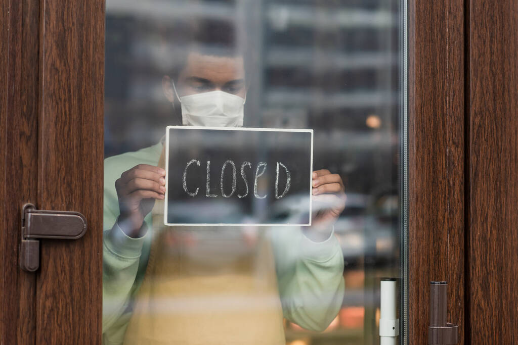 医療用マスクのアフリカ系アメリカ人バリスタで、カフェの入り口近くに閉じたレタリングが立っている黒板  - 写真・画像