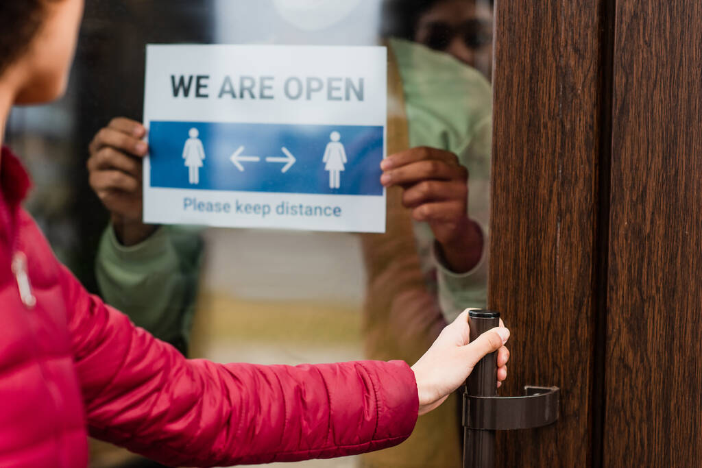Ausgeschnittene Ansicht des Kunden öffnende Tür in der Nähe afrikanisch-amerikanischen Barista mit offenen Schriftzug auf dem Schild in Café  - Foto, Bild