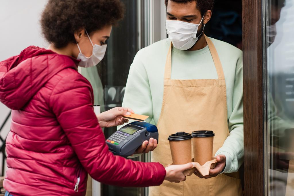 Barista afroamericano en máscara médica sosteniendo café y terminal de pago cerca del cliente con tarjeta de crédito  - Foto, imagen
