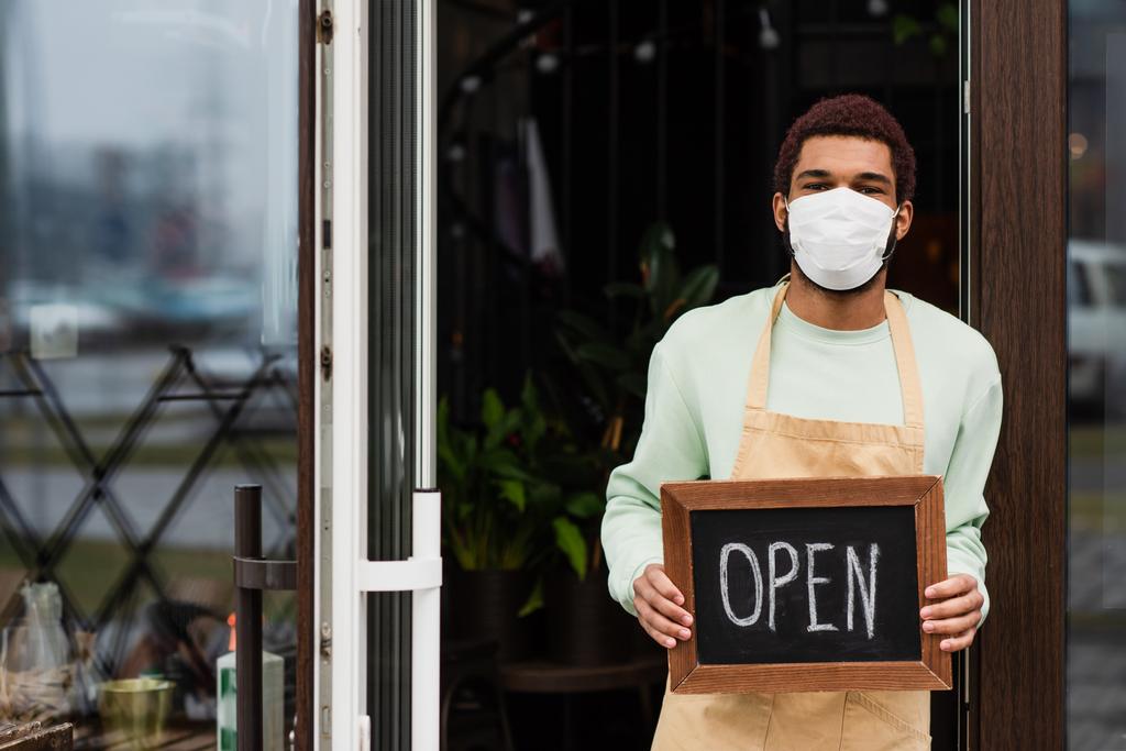 Barista afro-américaine en masque médical tenant un tableau noir avec lettrage ouvert près de la porte du café  - Photo, image