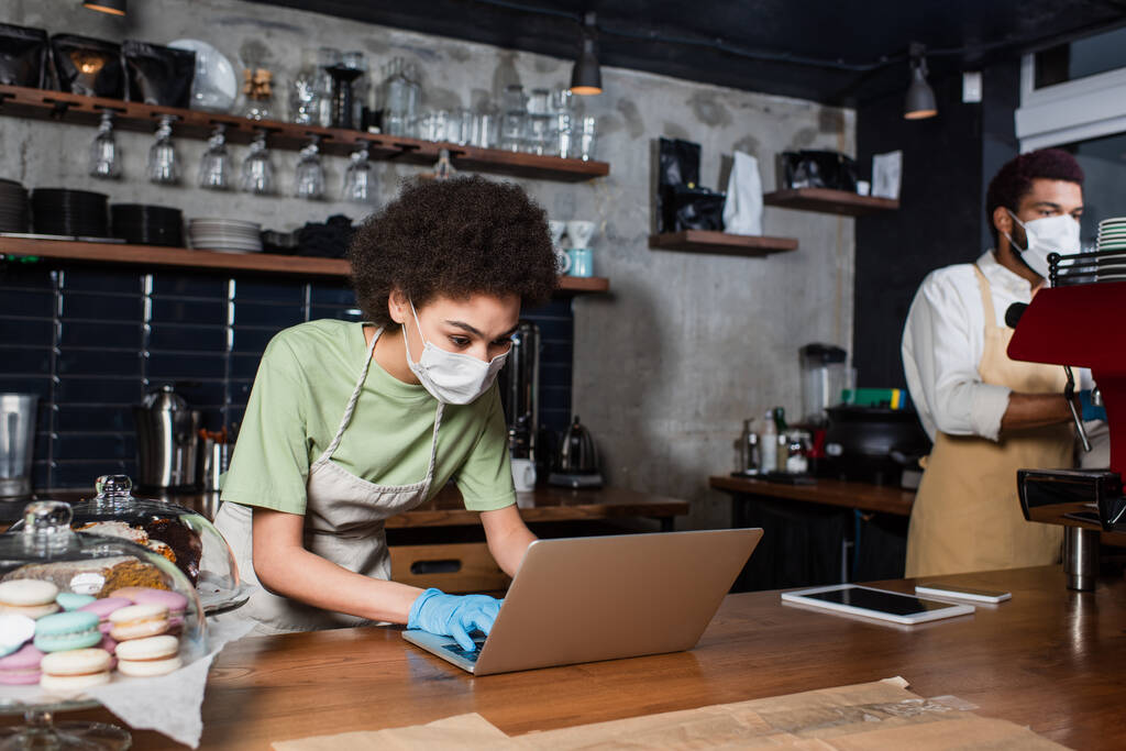 Африканський барист у медичній масці за допомогою ноутбука біля розмитого колеги та кавоварки у кафе.  - Фото, зображення