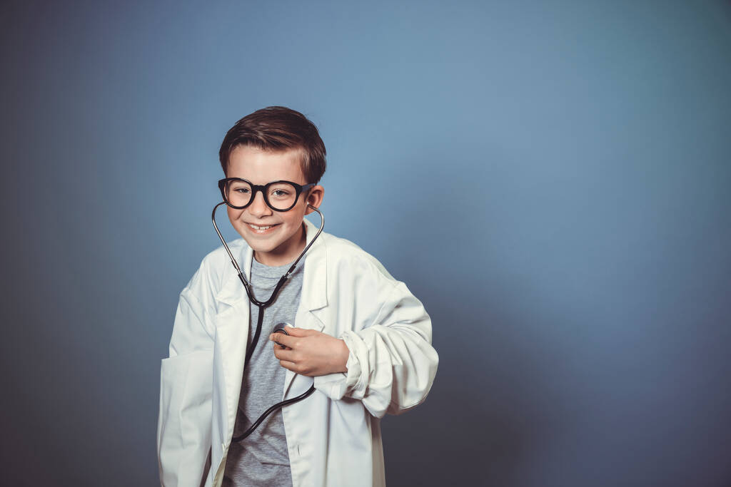 クールな少年は白い医者のスモックを持つ医者として偽装されており、青い背景の前で注射器と聴診器で遊ぶ - 写真・画像