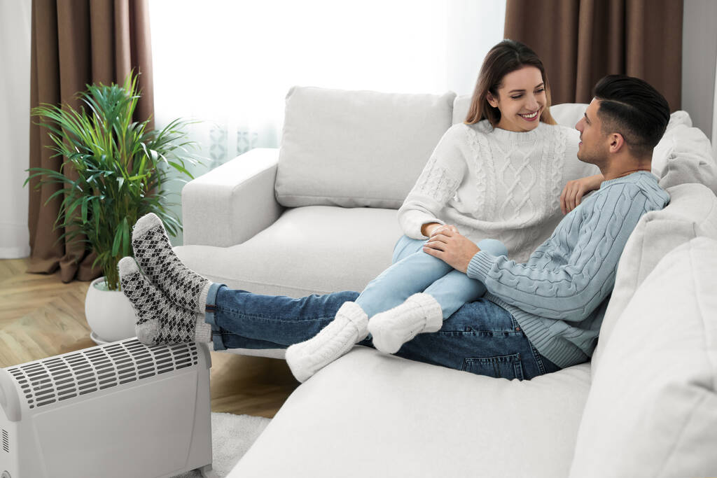 Ευτυχισμένο ζευγάρι κάθεται σε καναπέ κοντά σε ηλεκτρικό θερμαντήρα στο σπίτι - Φωτογραφία, εικόνα