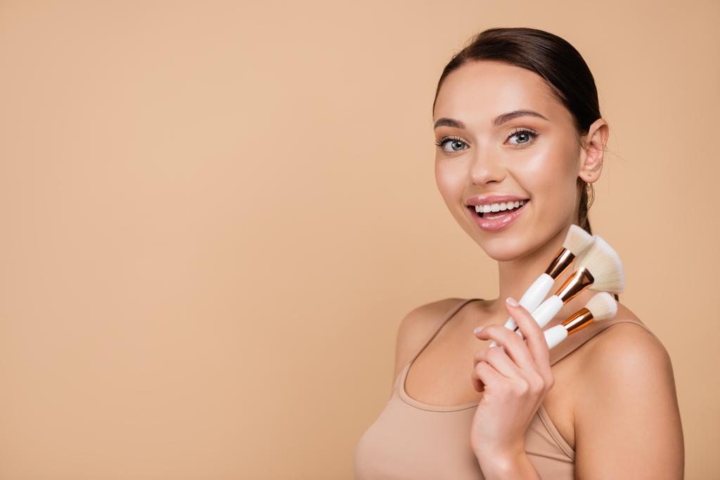 joyful young woman with cosmetic brushes looking at camera isolated on beige - Photo, Image