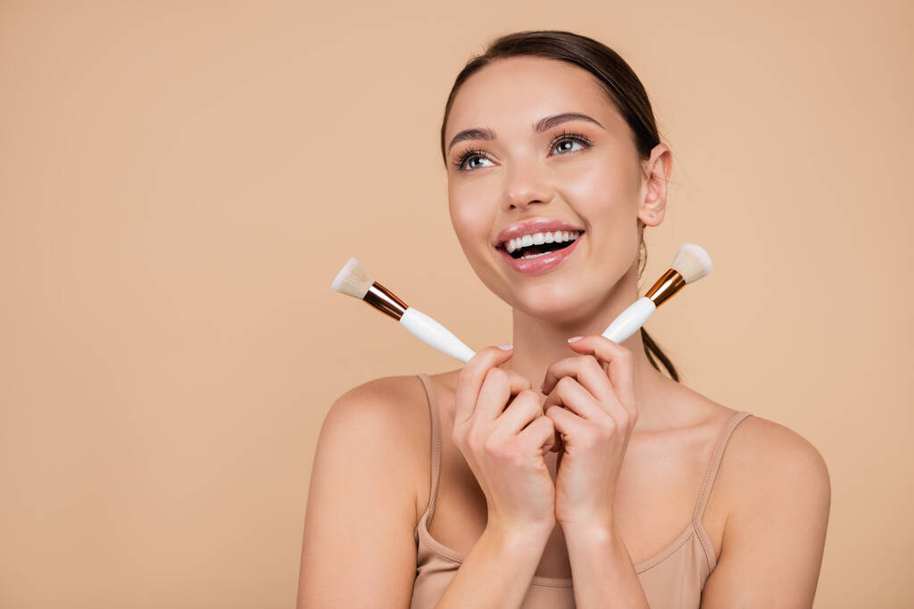 excited woman with cosmetic brushes looking away isolated on beige - Photo, Image