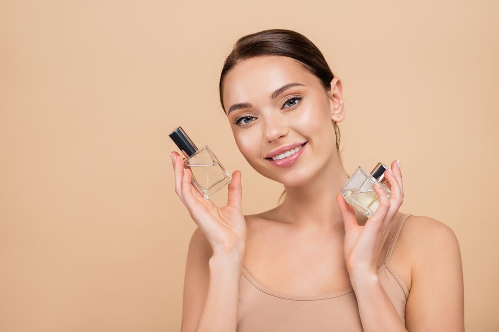 mujer joven con frascos de perfume sonriendo a la cámara aislada en beige - Foto, imagen