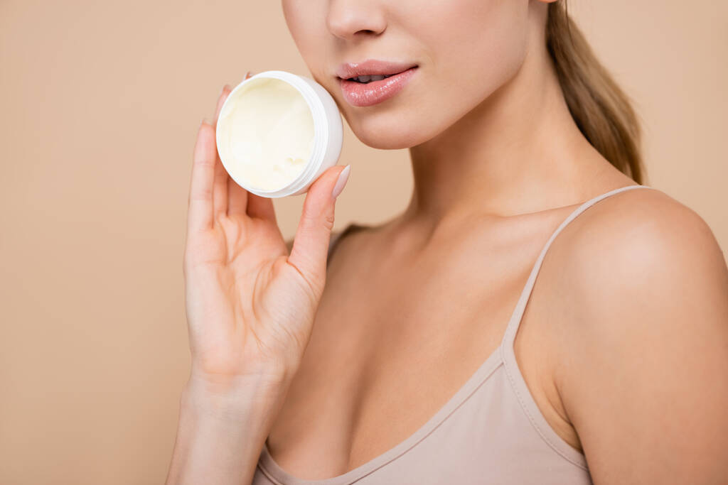 cropped view of woman with shiny lips holding cosmetic cream isolated on beige - Photo, Image