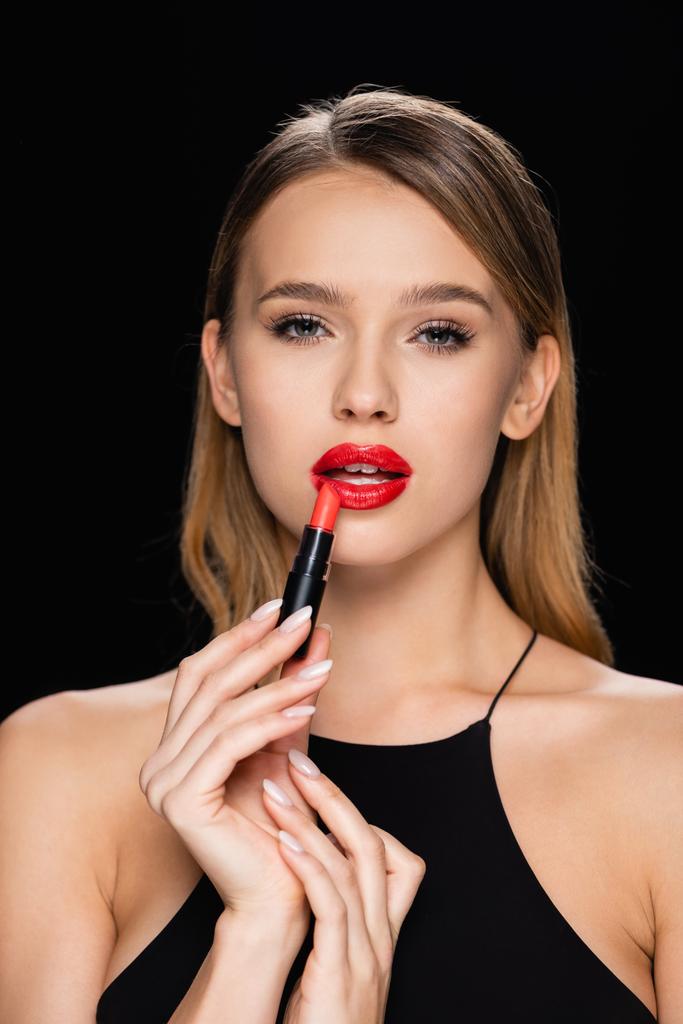 charming woman looking at camera while applying red lipstick isolated on black - Photo, Image
