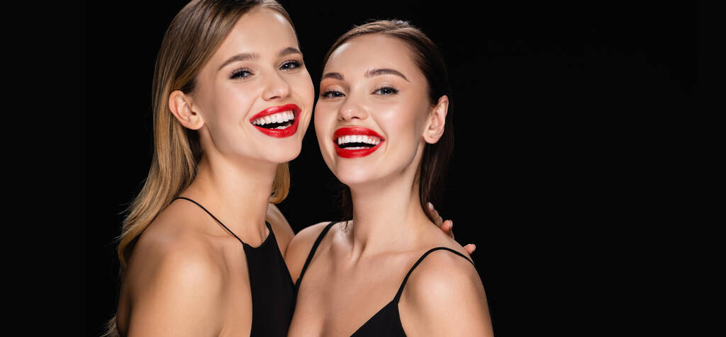 dos mujeres encantadoras con maquillaje brillante riendo de la cámara aislada en negro, pancarta - Foto, imagen