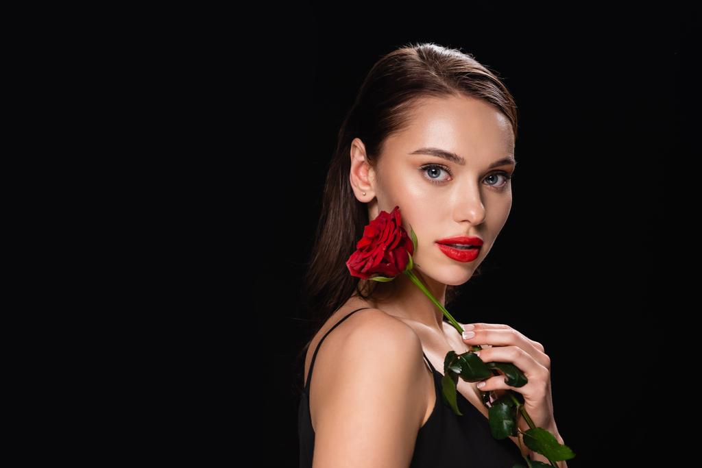 mooie vrouw met rode lippen en roos kijken naar camera geïsoleerd op zwart - Foto, afbeelding