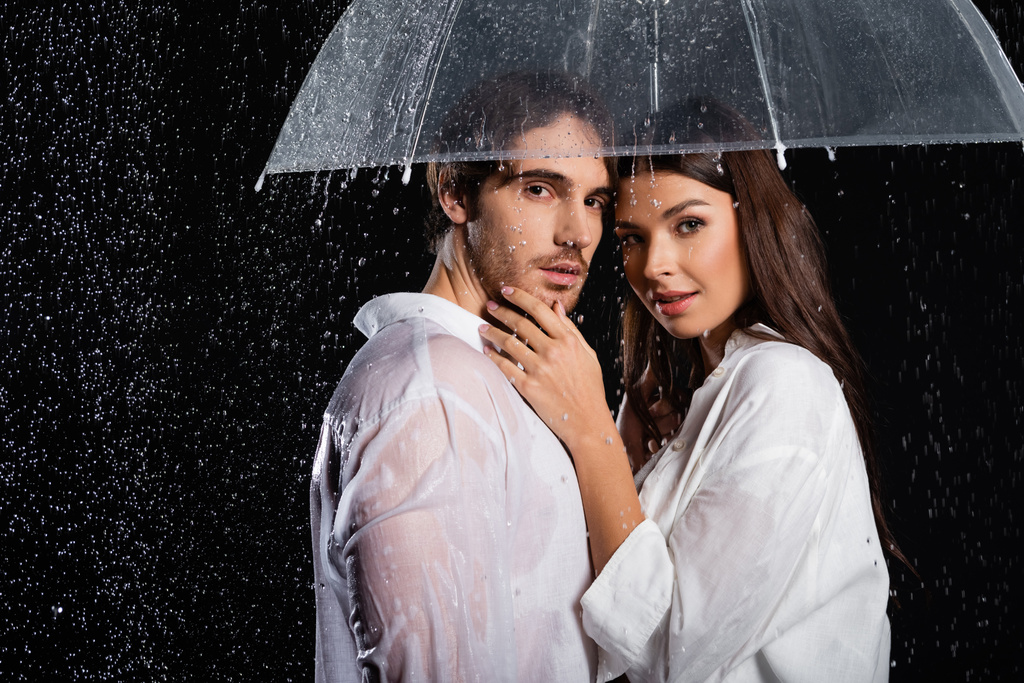 romantikus fiatal felnőtt pár áll esőben esernyővel és nézi kamera fekete háttér - Fotó, kép