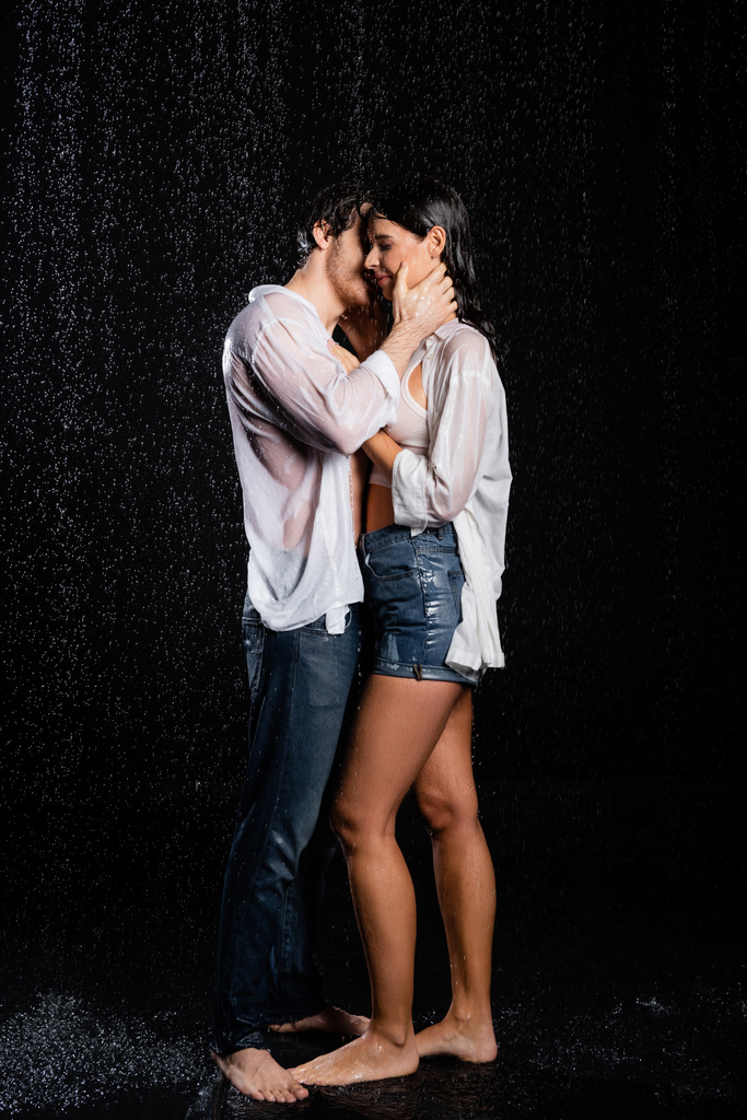 романтическая молодая мокрая пара, стоящая под дождем и чувственно обнимающая руками на черном фоне - Фото, изображение