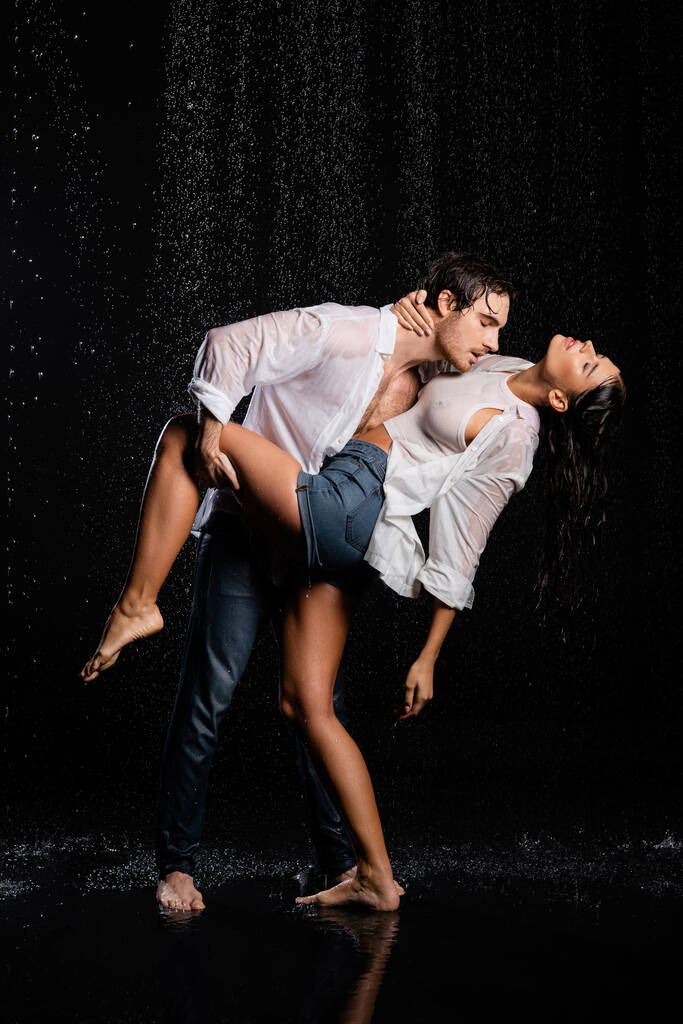 mokrý dobře vypadající muž vášnivě objímající ženu a drží její nohu po ruce v dešti kapky na černém pozadí - Fotografie, Obrázek