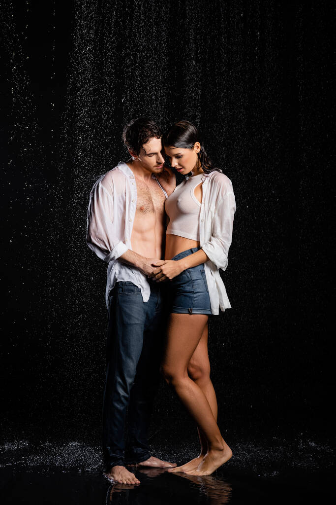 natte knappe man zachtjes knuffelen en vasthouden van handen met mooie vrouw in regen druppels op zwarte achtergrond - Foto, afbeelding