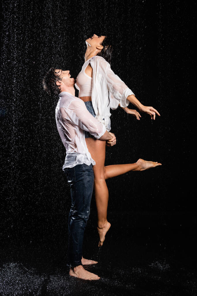 glücklich Freund heben lächelnde Freundin in nassen Kleidern in Regentropfen auf schwarzem Hintergrund - Foto, Bild