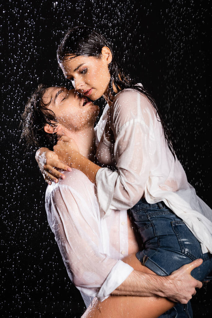 couple romantique sexy en chemises blanches mouillées serrant passionnément dans les gouttes de pluie sur fond noir - Photo, image