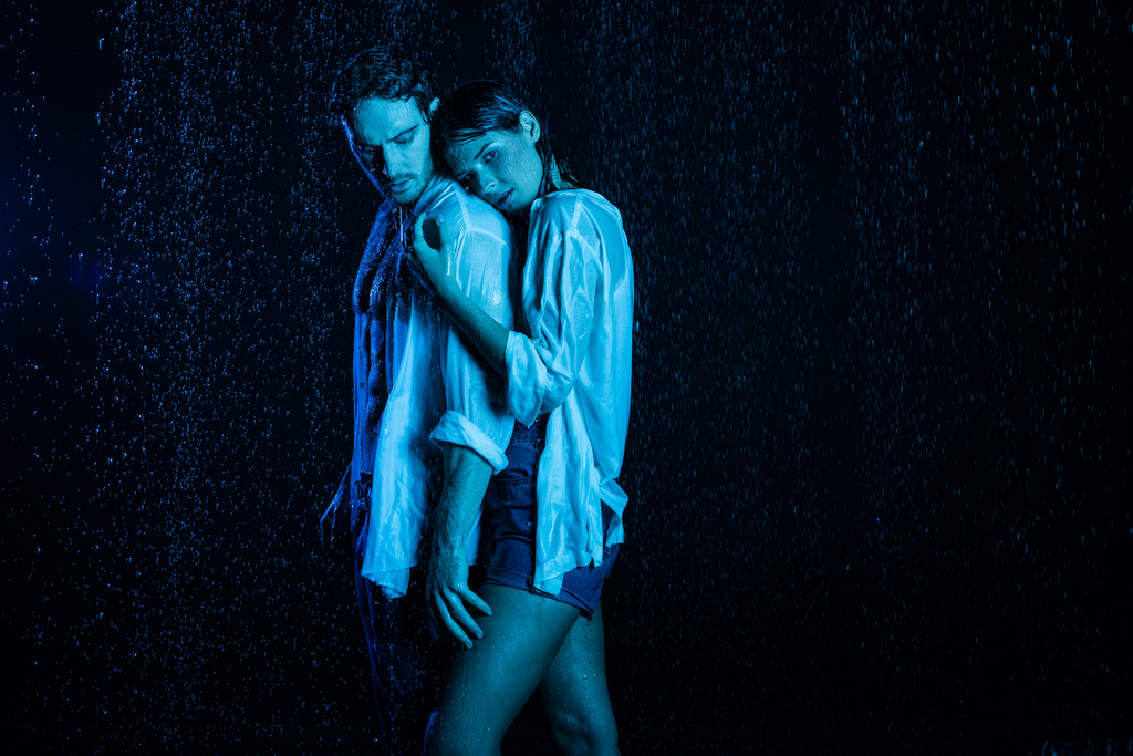nasses romantisches Paar, das sich sanft in Wassertropfen auf schwarzem Hintergrund mit blauem Farbfilter umarmt  - Foto, Bild