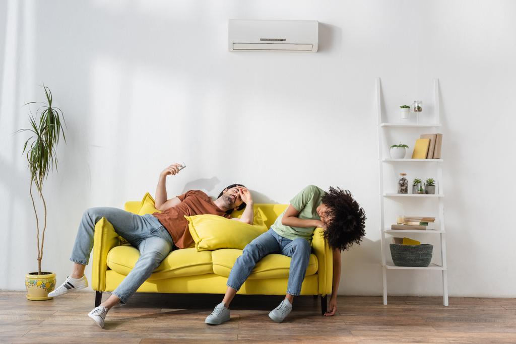 межрасовая пара, страдающая от жары на желтом диване в современной гостиной  - Фото, изображение