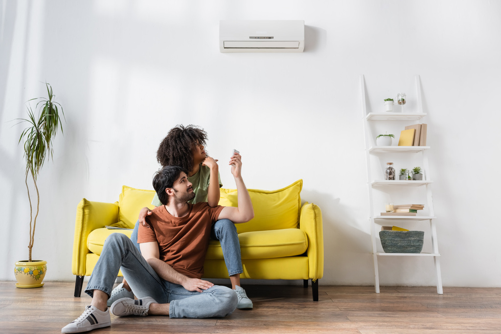 displeased interracial couple looking at air conditioner while sitting on sofa  - Photo, Image