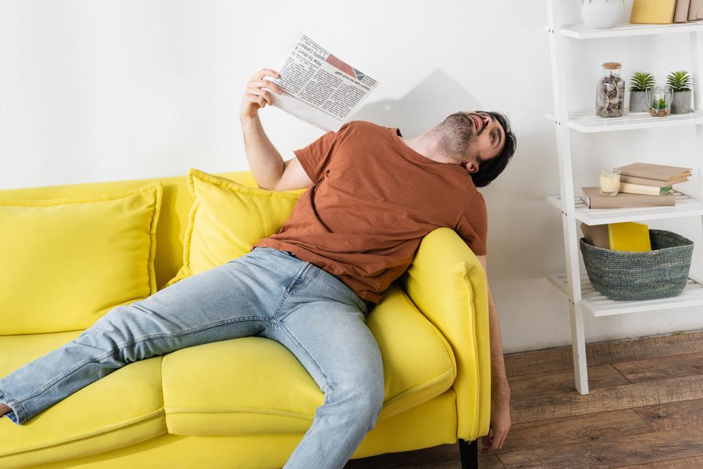 Mann fuchtelt mit Zeitung auf gelbem Sofa und leidet unter Hitze - Foto, Bild
