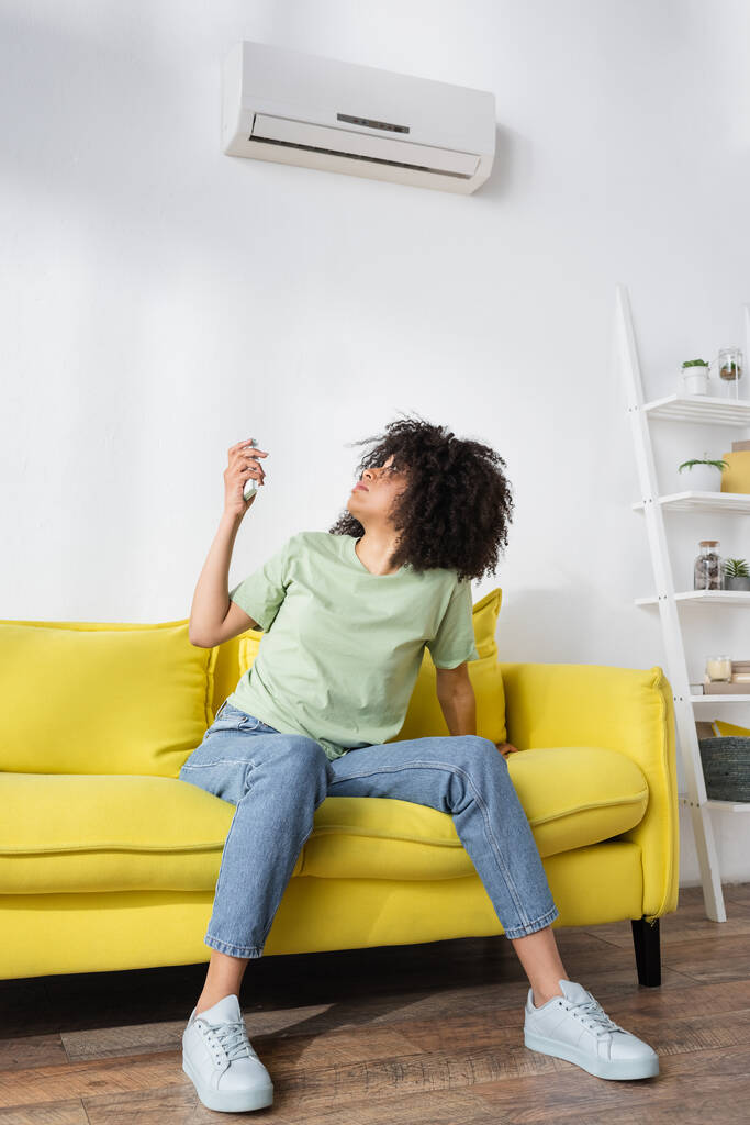 donna afro-americana delusa che tiene il telecomando mentre siede sul divano giallo e soffre di calore - Foto, immagini