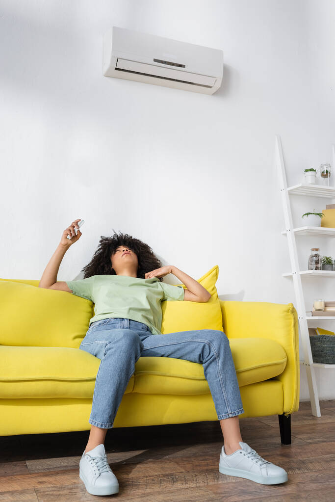 donna afro-americana scontenta che tiene in mano il telecomando mentre è seduta sul divano giallo e soffre di calore - Foto, immagini