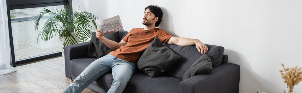 Mann winkt mit Zeitung, während er auf grauem Sofa liegt und unter Hitze leidet - Foto, Bild