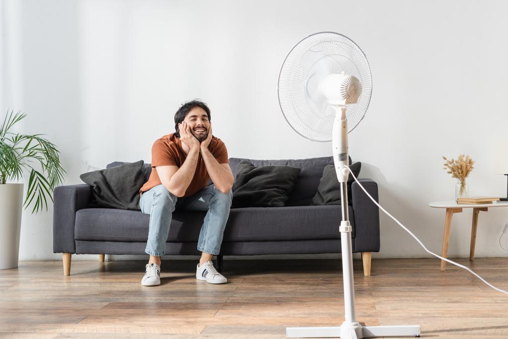 щасливий бородатий чоловік сидить на дивані біля розмитого електричного вентилятора
  - Фото, зображення