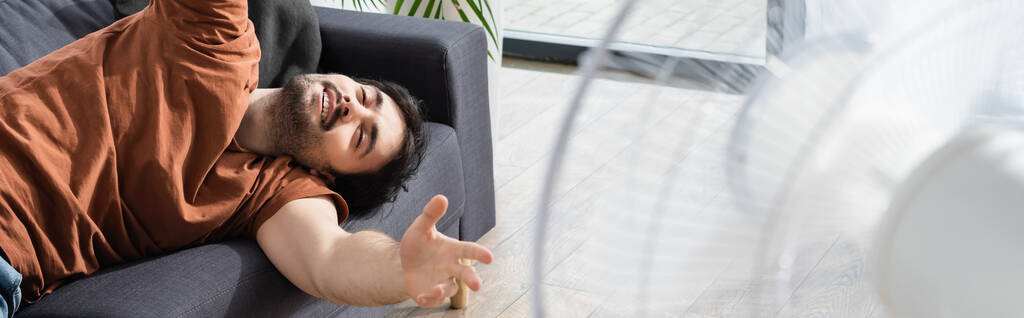 jovem satisfeito deitado no sofá perto de ventilador elétrico borrado, banner - Foto, Imagem