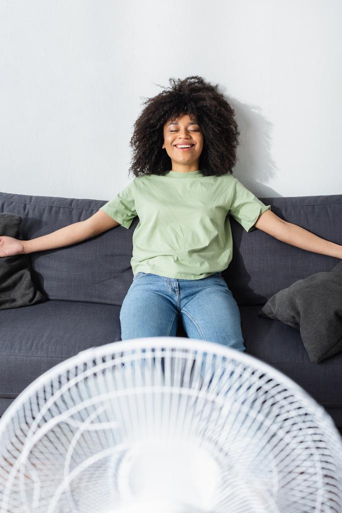 joyeuse femme afro-américaine reposant sur un canapé près d'un ventilateur électrique flou - Photo, image