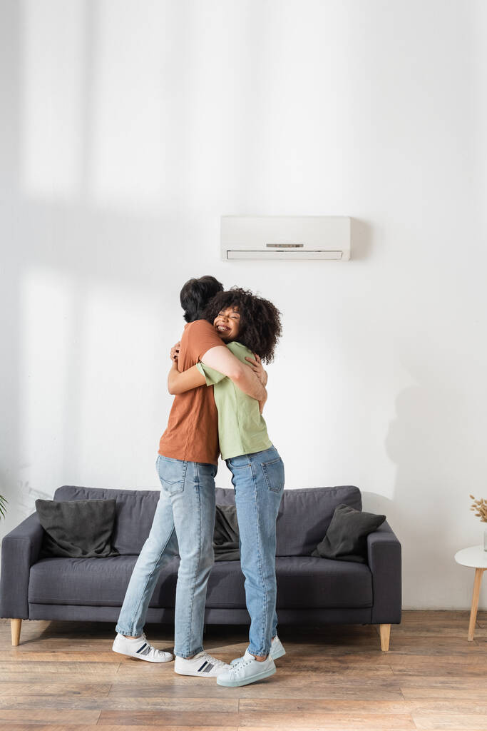abrazo multiétnico feliz cerca de aire acondicionado fijo en la sala de estar moderna - Foto, imagen