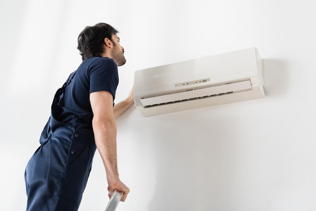 Handwerker in Overalls steht auf Leiter, während er kaputte Klimaanlage repariert  - Foto, Bild