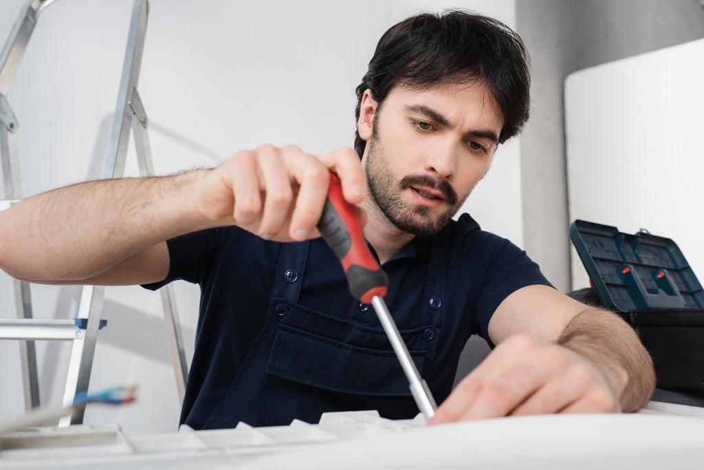 Konzentrierter Heimwerker in Overalls hält Schraubenzieher während er kaputte Klimaanlage repariert  - Foto, Bild